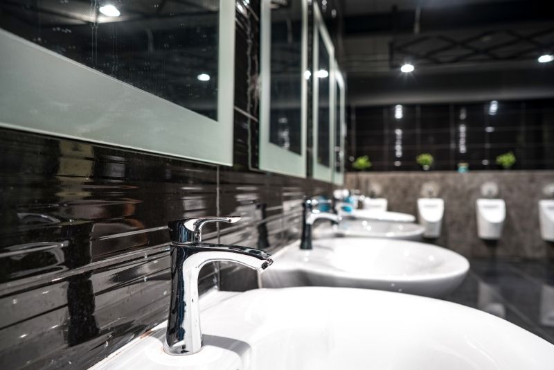 Dark-tiled commercial restroom with chrome tapware over a row of sinks and urinals in the background