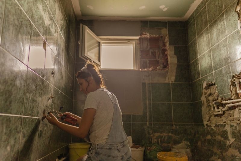 Tradesperson retiling a green bathroom wall during a renovation, with the window opened up to exposed brickwork